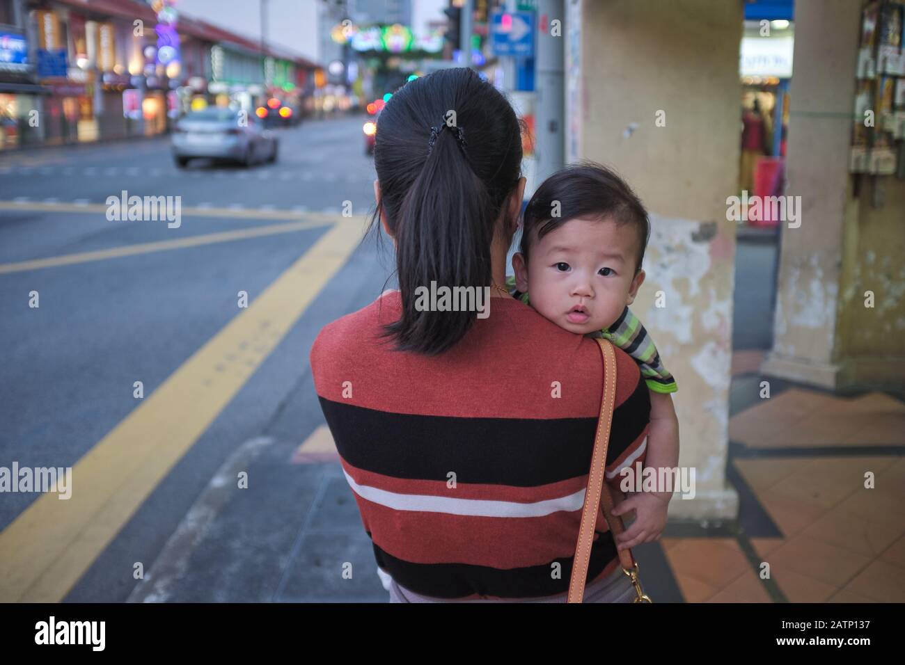 Eine Chinesin aus Singapur, die den jungen Sohn auf der Schulter trägt, der gerade und neugierig in die Kamera blickt Stockfoto