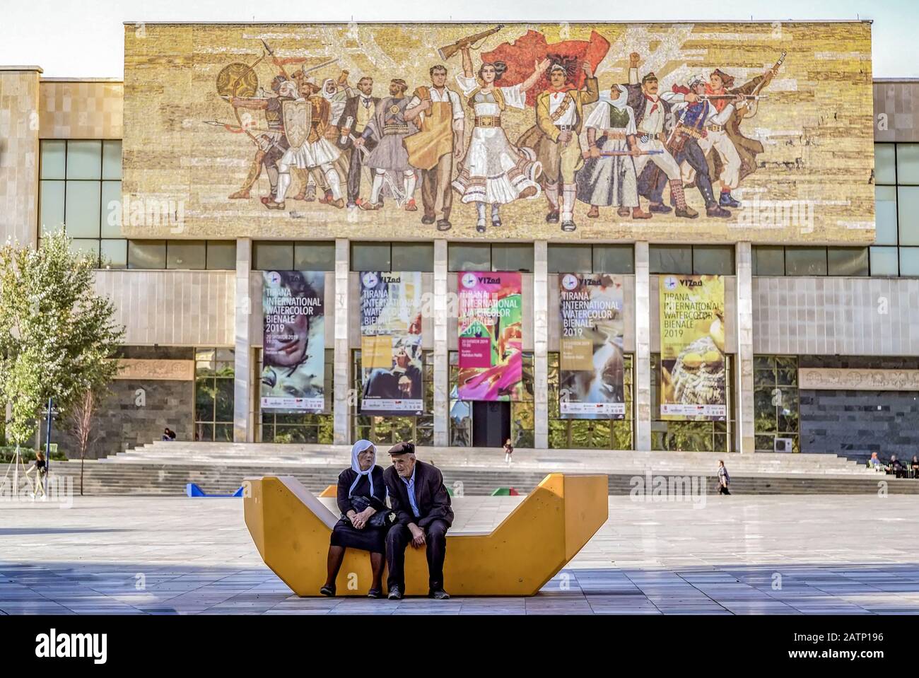 Älteres albanisches Paar sitzt auf dem Skanderbeg-Platz vor dem nationalen Geschichtsmuseum.Mosaik zeigt die siegreichen Albaner aus der Geschichte. Stockfoto