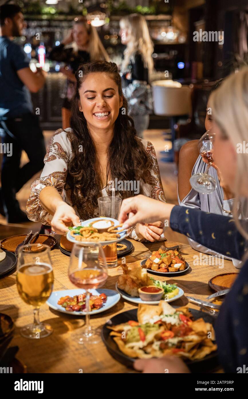 Eine Gruppe von Freunden bei einer Mahlzeit zusammen, der Hauptfokus ist eine Frau, die einen Teller Essen heraushält, damit eine andere Frau von ihr auswählen kann. Stockfoto