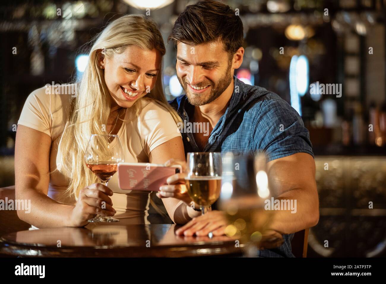 Ein heterosexuelles Paar sitzt und trinkt zusammen in einer Bar, die das Telefon der Frau beim Lächeln betrachtet. Stockfoto