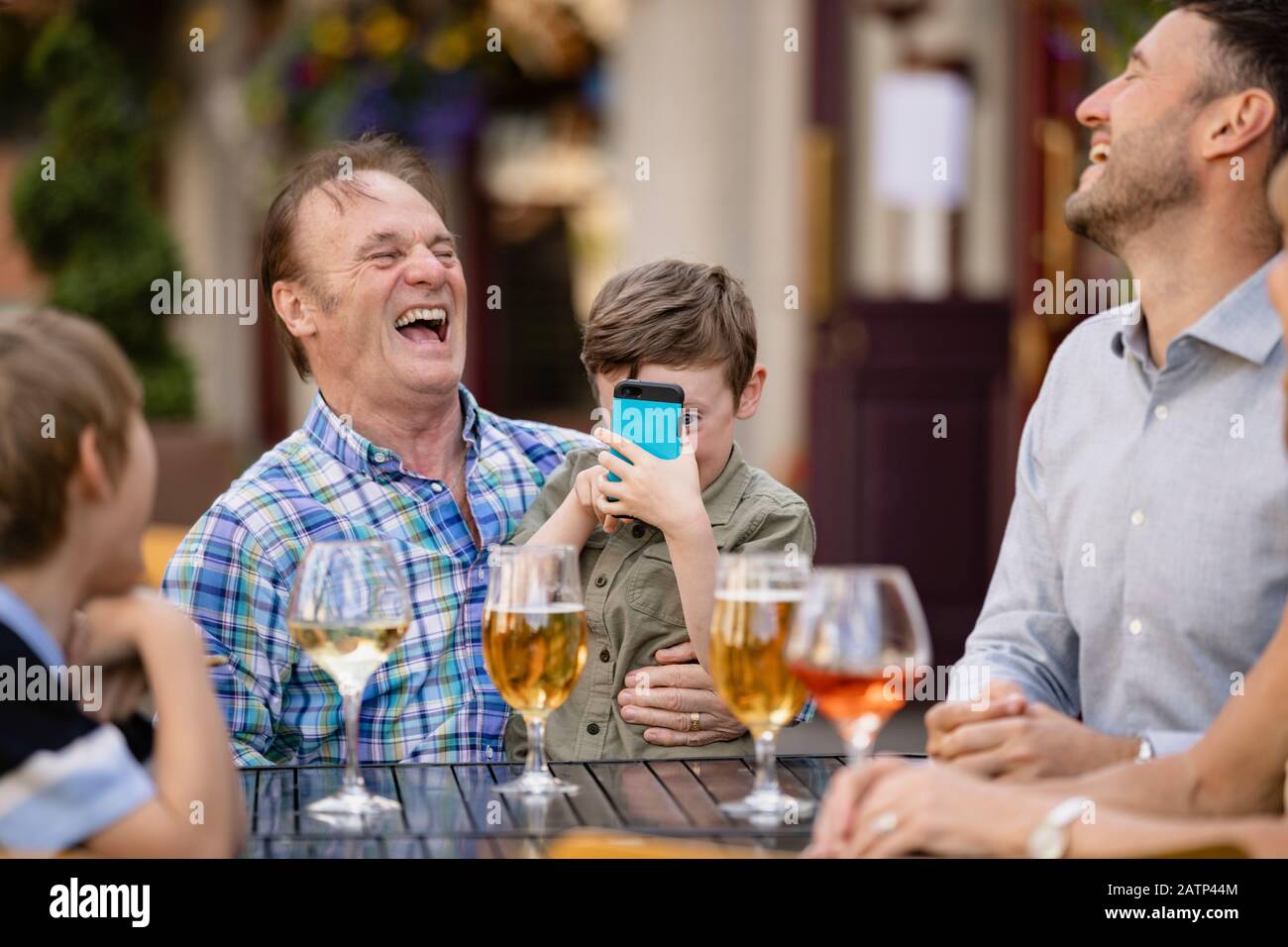 Eine mehrköpfige Familie lacht zusammen, während sie im Freien in einem städtischen Biergarten trinkt. Im Mittelpunkt steht ein kleiner Junge, der auf seinem Großfath sitzt Stockfoto