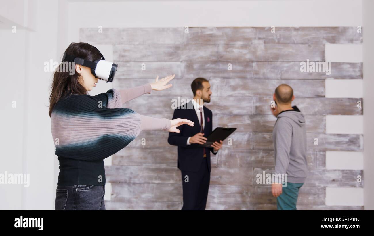 Designerin mit virtueller Reality-Brille in leerer Wohnung, während Immobilienmakler im Hintergrund mit dem Hausbesitzer spricht. Stockfoto