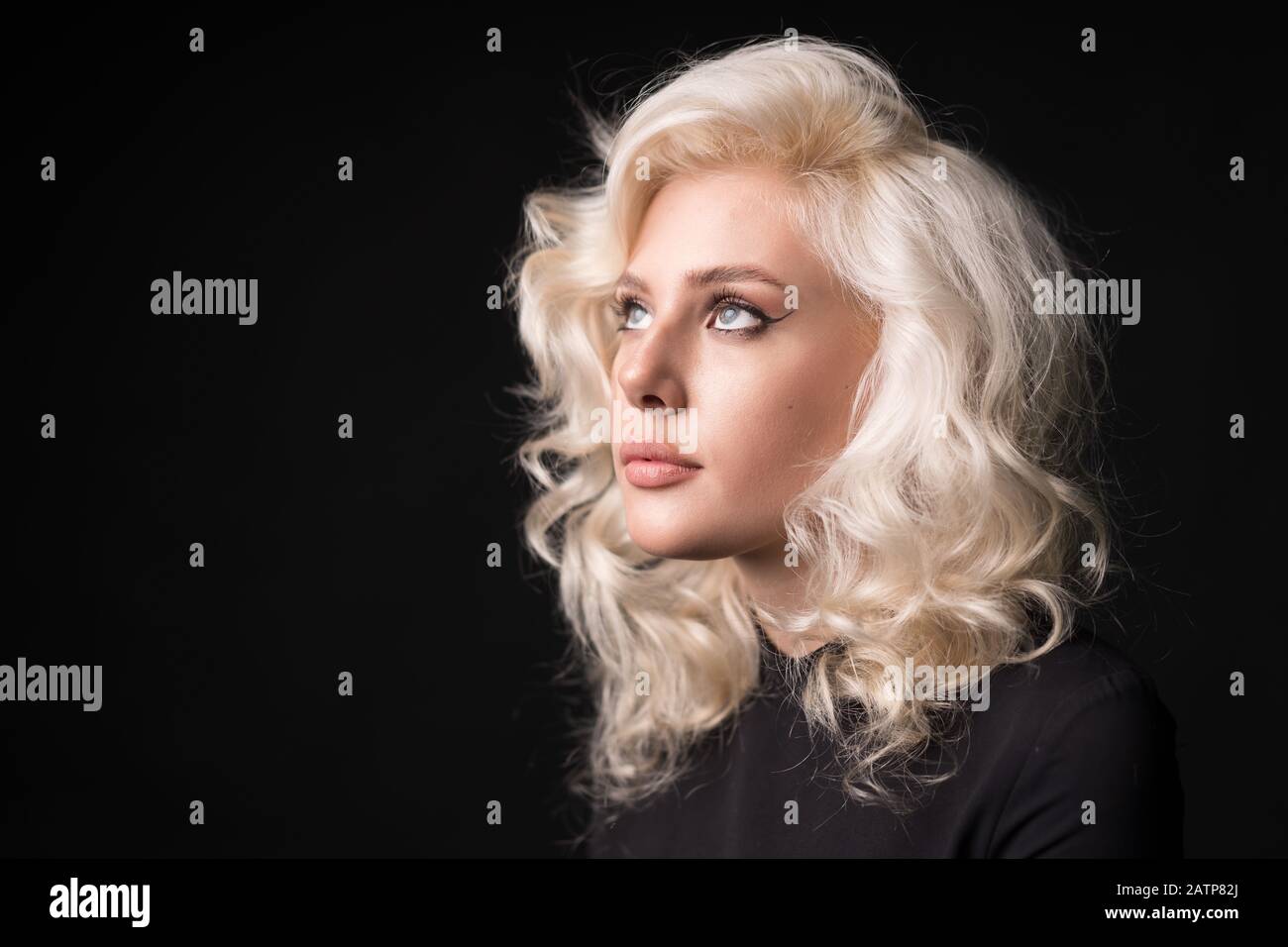 Charmantes blondes lockiges Mädchen, das isoliert auf schwarzem Hintergrund aufblickt. Stockfoto