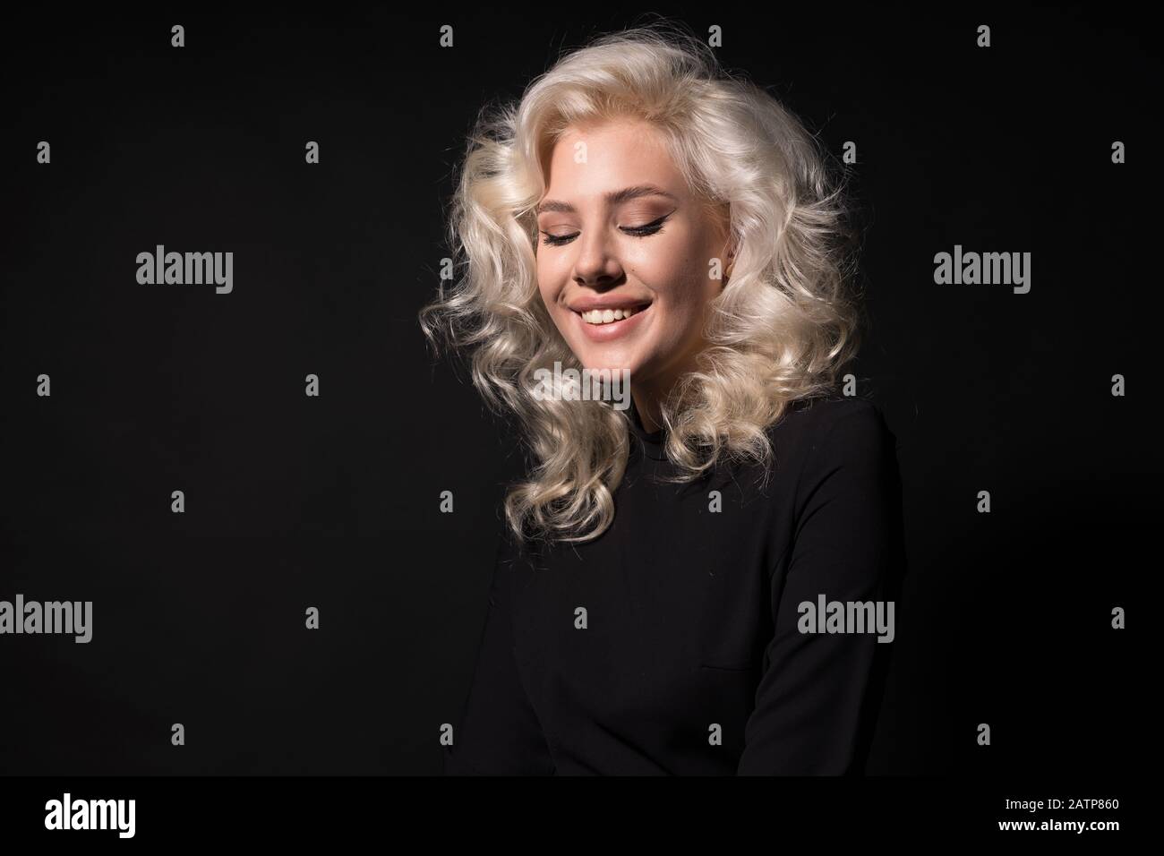 Blonde junge Frau lächelt mit geschlossenen Augen isoliert auf schwarzem Hintergrund. Stockfoto