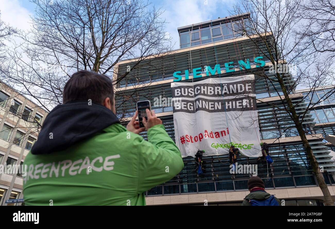 München, Deutschland. Februar 2020. Seil dich an Greenpeace-Aktivisten hängen an der façade des Siemens-Hauptquartiers in der Innenstadt vor einem Banner, das sagt: "Bush-Brände beginnen hier! - #StopAdani', während ein Mann in einer Greenpeace-Jacke sein Handy auf die Aktion hinweist. Einen Tag vor der Siemens-AGM protestiert die Umweltorganisation gegen den Beitrag des Industriekonzerns zu einem riesigen Kohleminen-Projekt der Adani-Gruppe in Australien. Kredit: Peter Kneffel / dpa / Alamy Live News Stockfoto