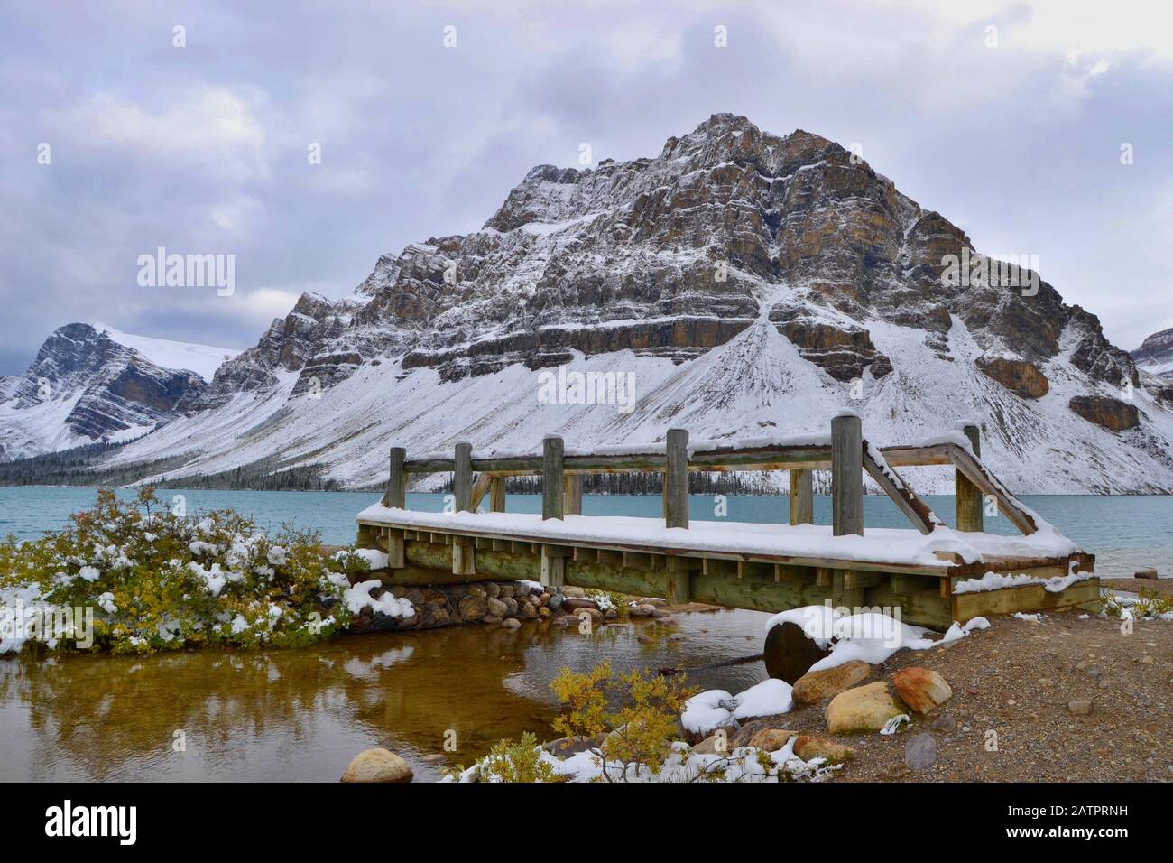 Blaues Wasser des Bow Lake, schneebedeckte Berge. Holzbrücke, graue Wolken, Blick vom Ufer. Banff National Park, Rocky Mountains, Kanada. Stockfoto