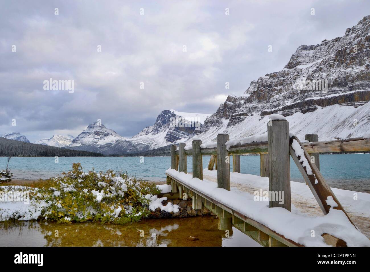 Blaues Wasser des Bow Lake, schneebedeckte Berge. Holzbrücke, graue Wolken, Blick vom Ufer. Banff National Park, Rocky Mountains, Kanada. Stockfoto