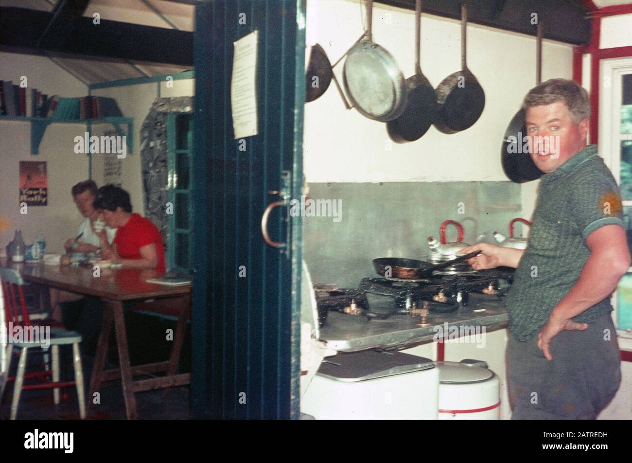 Ein Bild der Umgangssprache, das auf einer analogen 35-mm-Filmtransparenz aufgenommen wurde, glaubte, schwarze und silberne Bratpfanne auf dem Herd, mit dem Mann in der Verarbeitung des Kochens, in der Küche eines Restaurants oder einer Jugendherberge, 1970, darzustellen. Zu den Hauptthemen/Objekten, die erkannt wurden, gehört Restaurant. () Stockfoto
