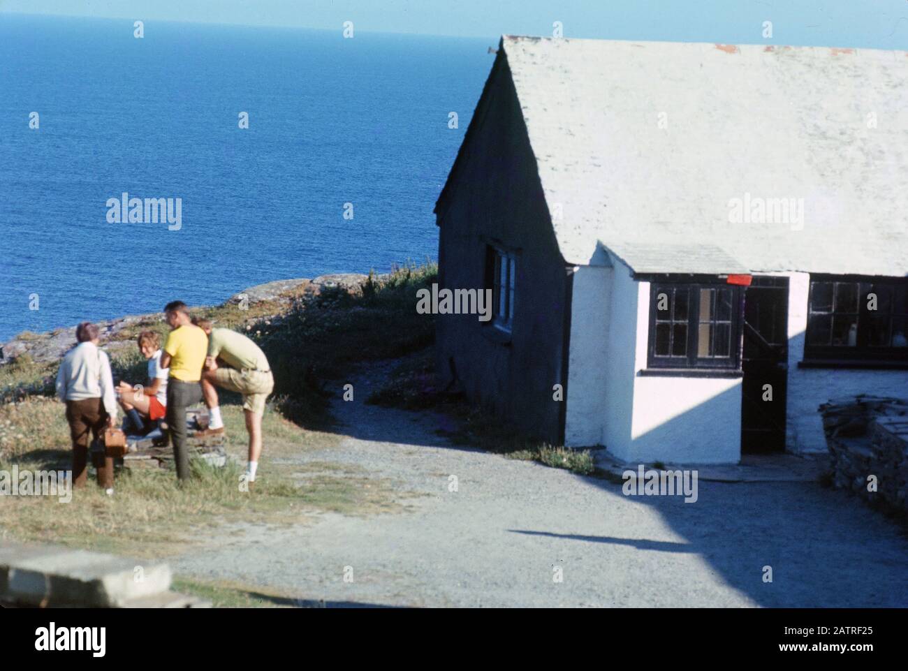 Ein Bild der Umgangssprache, das auf einer analogen 35-mm-Filmtransparenz aufgenommen wurde, glaubte, Frau in gelbem Hemd und blauen Denim-Shorts neben weißem und schwarzem Haus in der Nähe des Körpers des Wassers, 1970, darzustellen. Zu den wichtigsten Themen/Objekten, die entdeckt wurden, gehören Meer und Strand. () Stockfoto