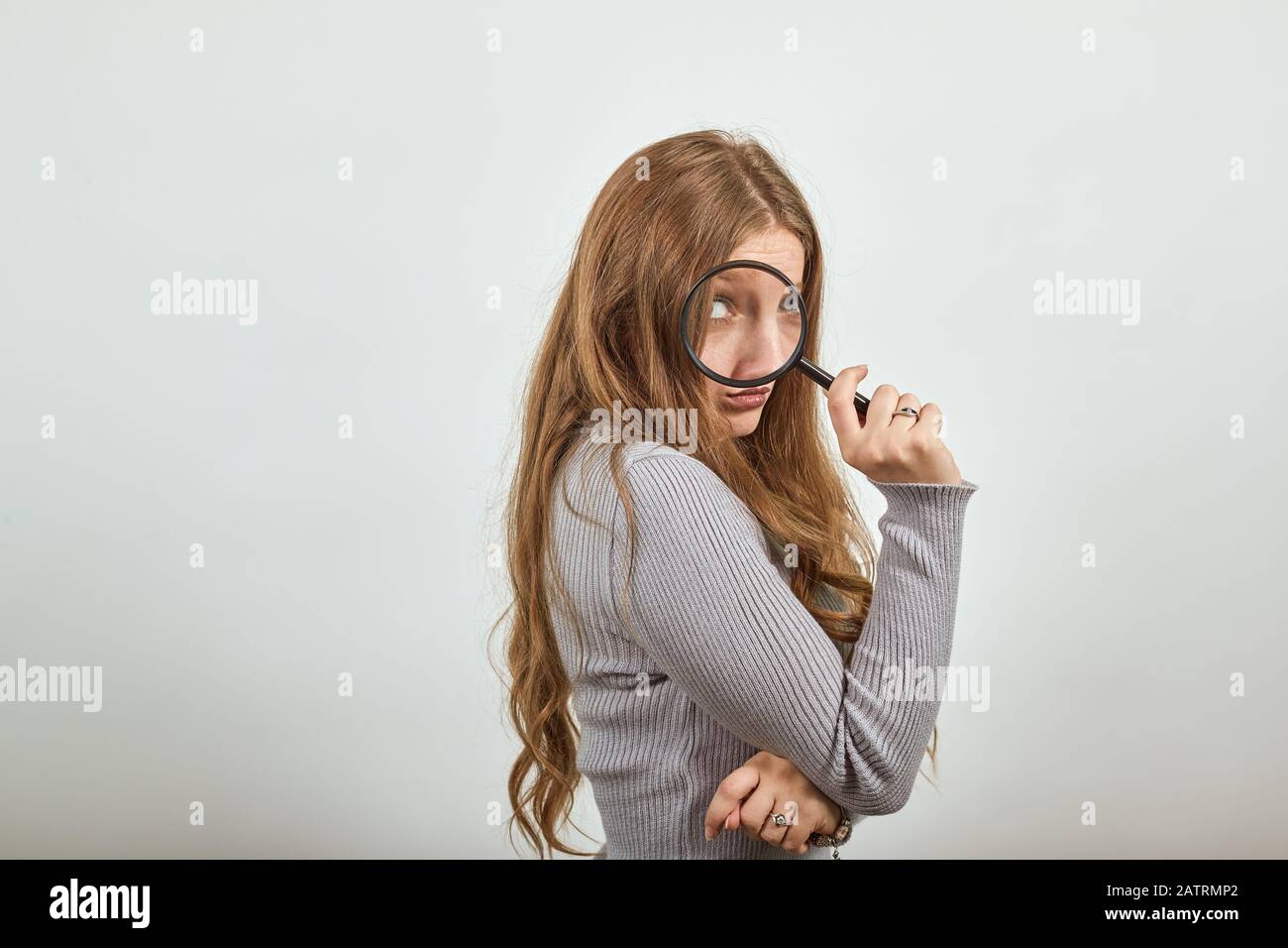 Frau in grauem Pullover schaut ein Auge größer als das andere durch Lupe Stockfoto