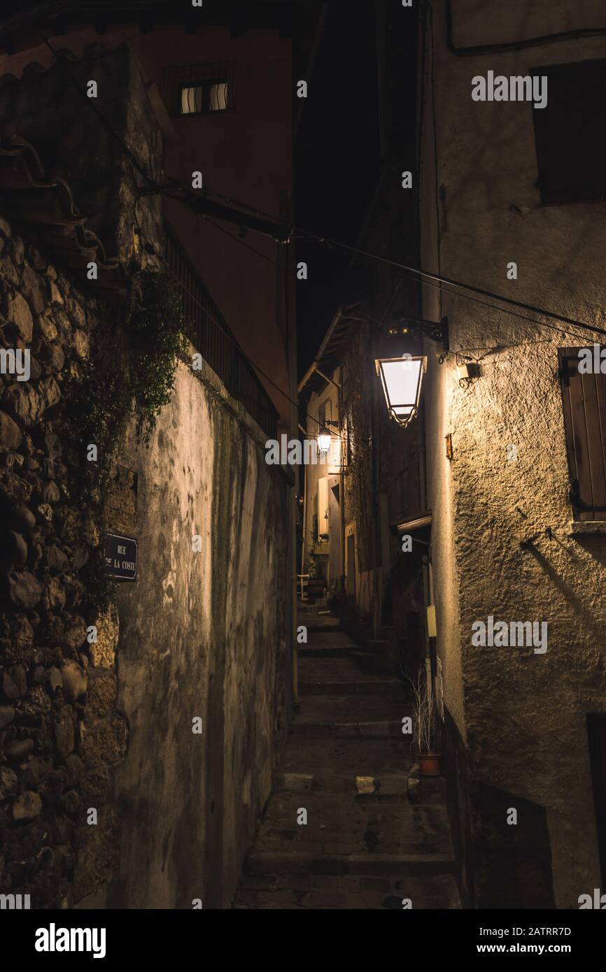 Eine mittelalterliche französische Gasse mit Treppen in der Nacht, die von Straßenbeleuchtungen beleuchtet wird (10. Januar 2020; gedreht an der Coste Street in Puget-Theniers, Frankreich) Stockfoto