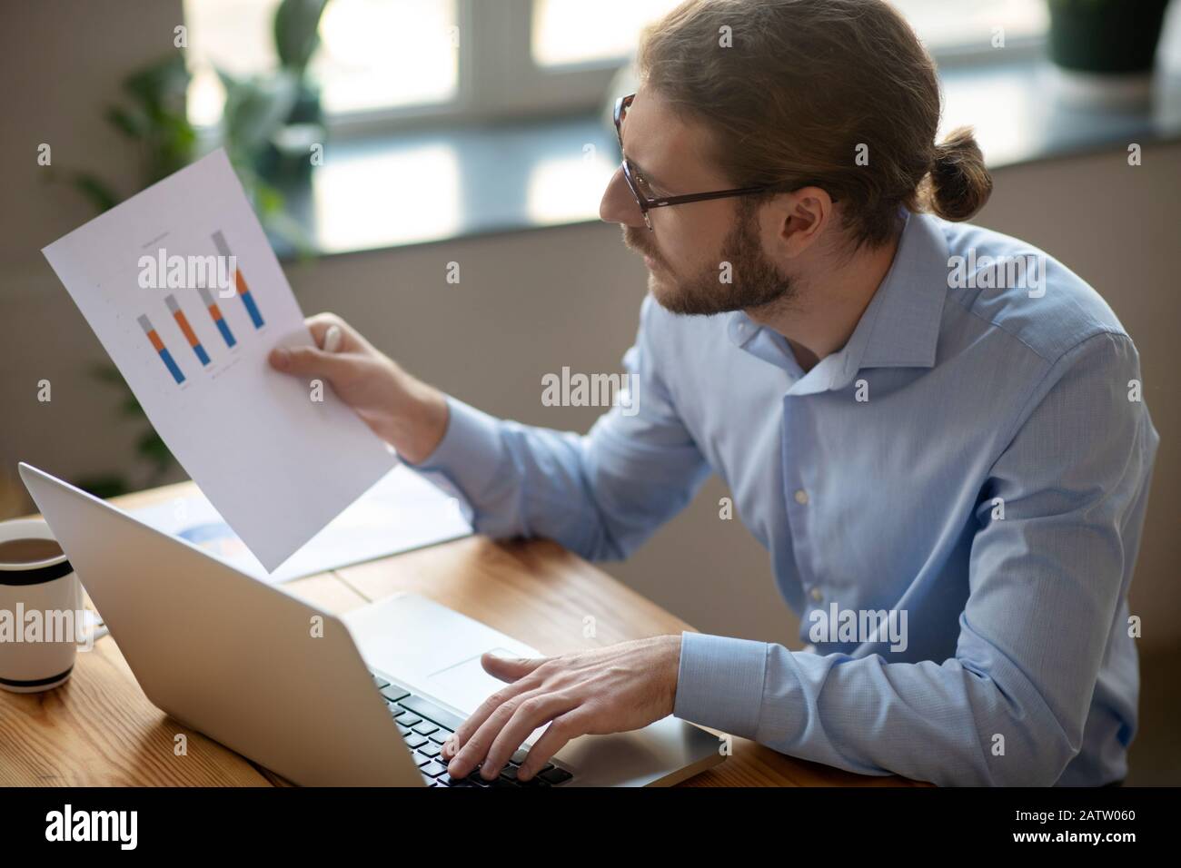 Durch Analyse der Daten. Ein ernsthafter junger Mann in einem hellblauen Hemd und einer Brille, der am Tisch sitzt und an einem Laptop arbeitet und vorsichtig auf den Schar blickt Stockfoto
