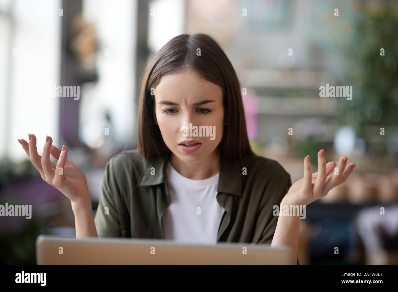 Unverständliche Situation. Müde Mädchen in schlechter Laune mit langen Haaren, die mit einer auffrostenden Augenbraue auf den Laptop-Bildschirm blicken und ihre Hände anheben. Stockfoto