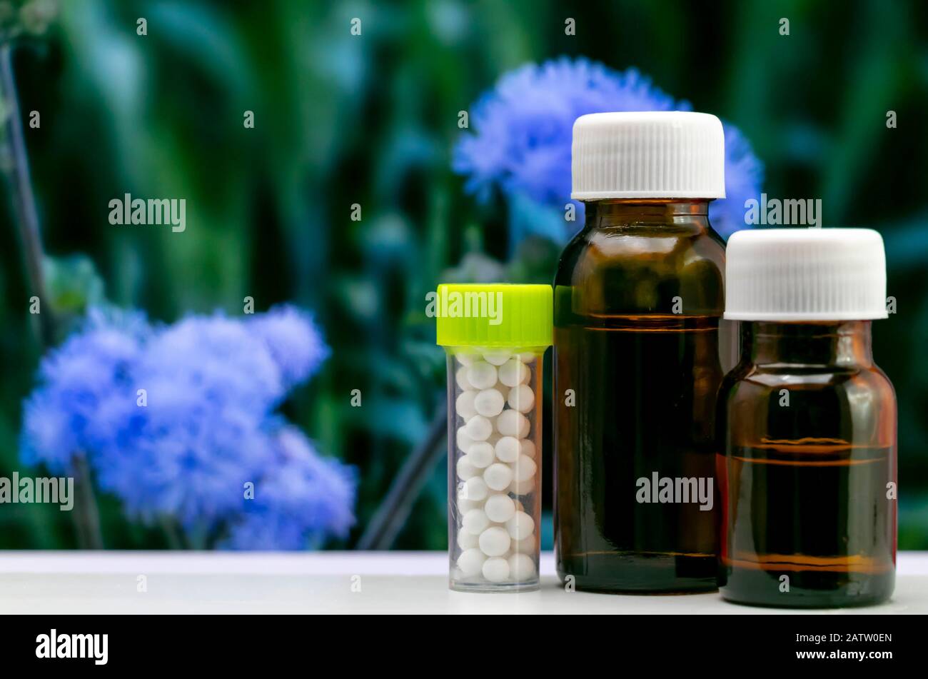 Modernes, Homöopathisches Konzept - Nahaufnahme der Homöopathischen Zuckerpillen und der flüssigen Substanz in einer Glasflasche mit verschwommenen blauen Blumen und grellen Stoffen Stockfoto