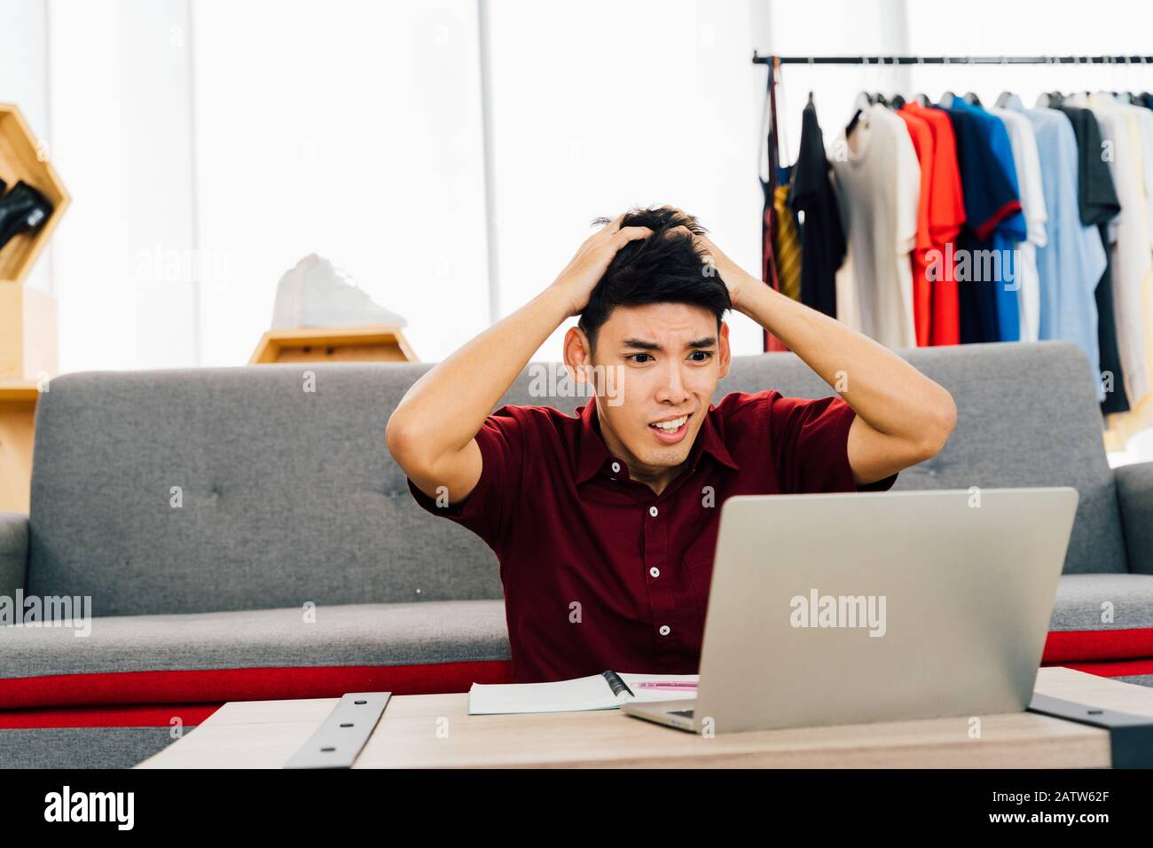 Schockierter junger Asiat in legerer Kleidung mit offenem Mund, der einen Bericht über das Projekt auf einem Laptop sieht, der zu Hause am Tisch sitzt Stockfoto