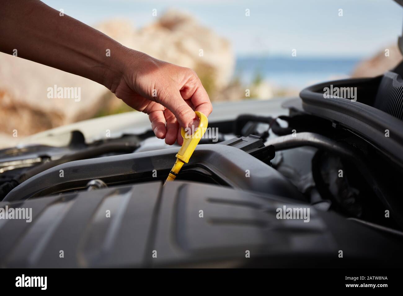 Motoröl prüfen. Die Hand einer Frau, die während einer Urlaubsreise ein Öl-Bajonett in einem modernen, beliebten Auto hält. Das Meer ist im Hintergrund sichtbar. Stockfoto