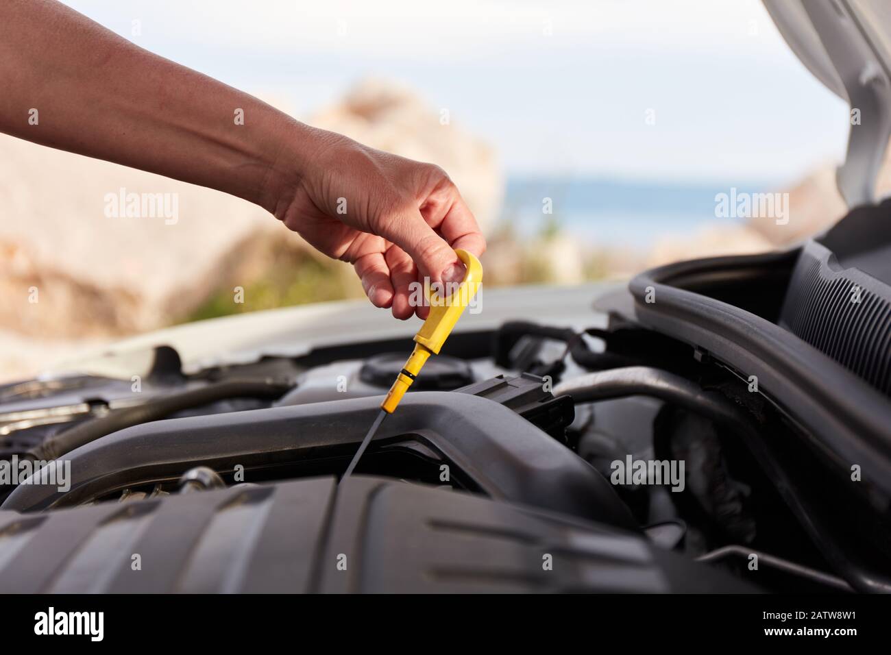 Motoröl prüfen. Die Hand einer Frau, die während einer Urlaubsreise ein Öl-Bajonett in einem modernen, beliebten Auto hält. Das Meer ist im Hintergrund sichtbar. Stockfoto
