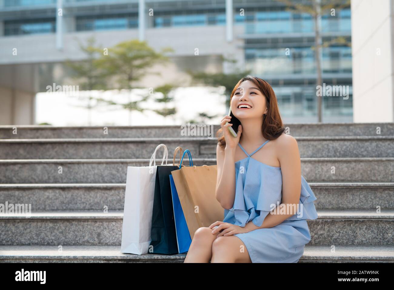 Fröhliche junge Asiatin mit Einkaufstaschen, die auf dem Handy telefonieren und auf einer Treppe sitzen. Stockfoto