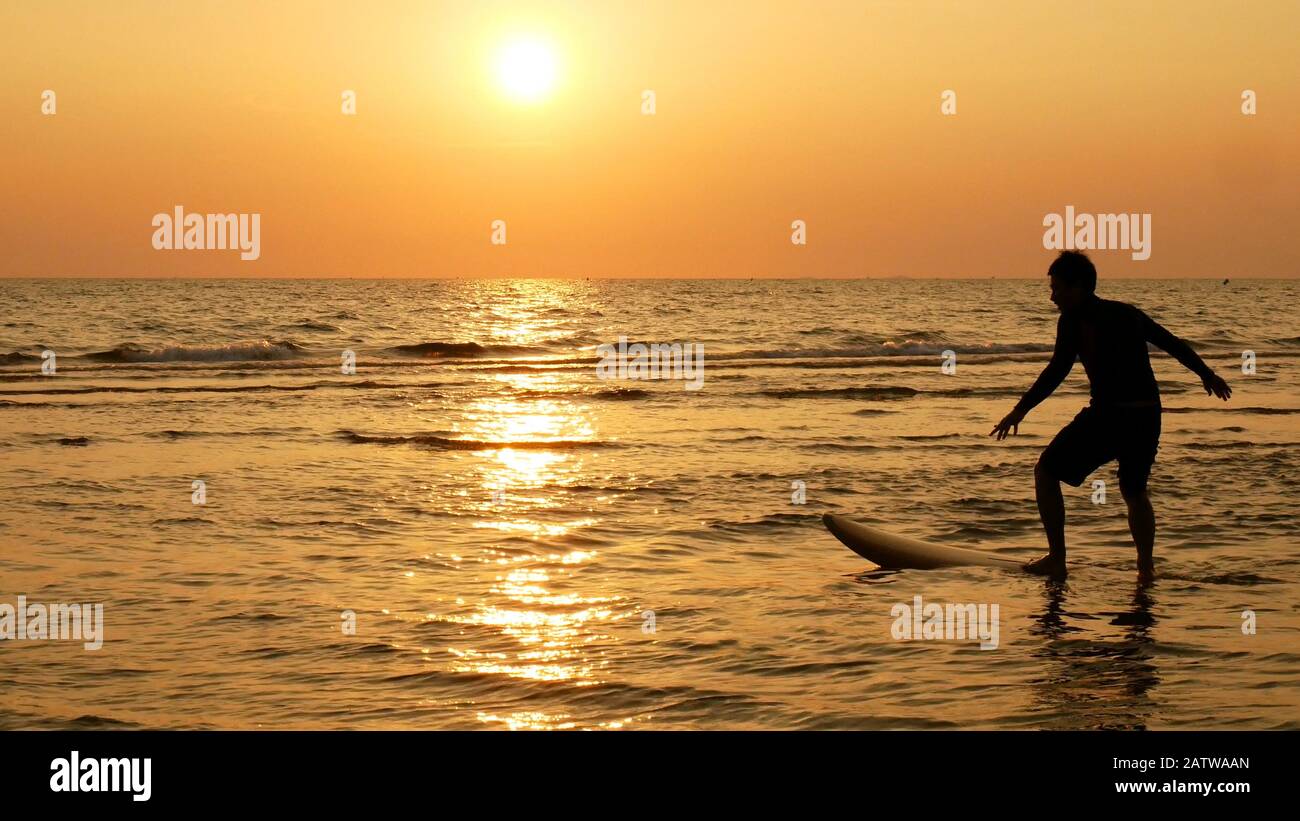 Silhouette des fröhlichen Surfmannes mit langen Surfbrettern bei Sonnenuntergang am tropischen Strand. Surfer am Strand an der Küste bei Sonnenuntergang mit schöner Aussicht Stockfoto