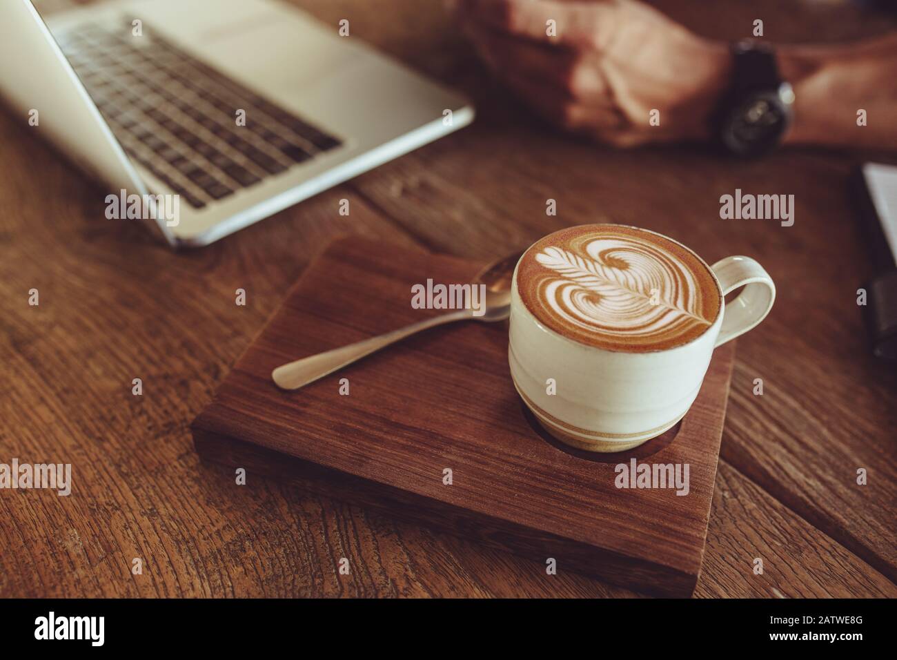 Nahaufnahme einer Tasse Cappuccino-Kunstkaffee auf dem Café-Tisch mit einem Mann, der am Laptop arbeitet. Kaffeetasse mit Kunstmuster aus Milchschaum. Stockfoto