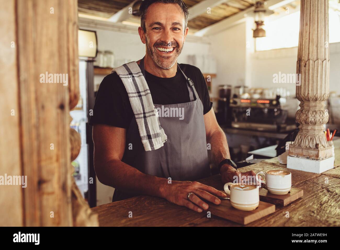 Barista männlich, der den Kunden Kaffee serviert. Ein mittelalter Erwachsener, der eine Schürze mit Blick auf die Kamera mit zwei Tassen Kaffee auf der Theke trägt. Stockfoto