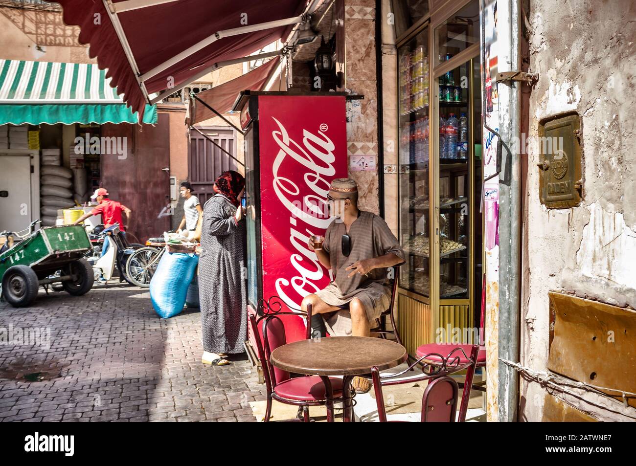 Marrakesch, Marokko. Oktober 2015. Der alte Mann sitzt in traditioneller marokkanischer Kleidung außerhalb eines Cafés, das Minztee neben einem modernen Coca Cola Machin trinkt Stockfoto