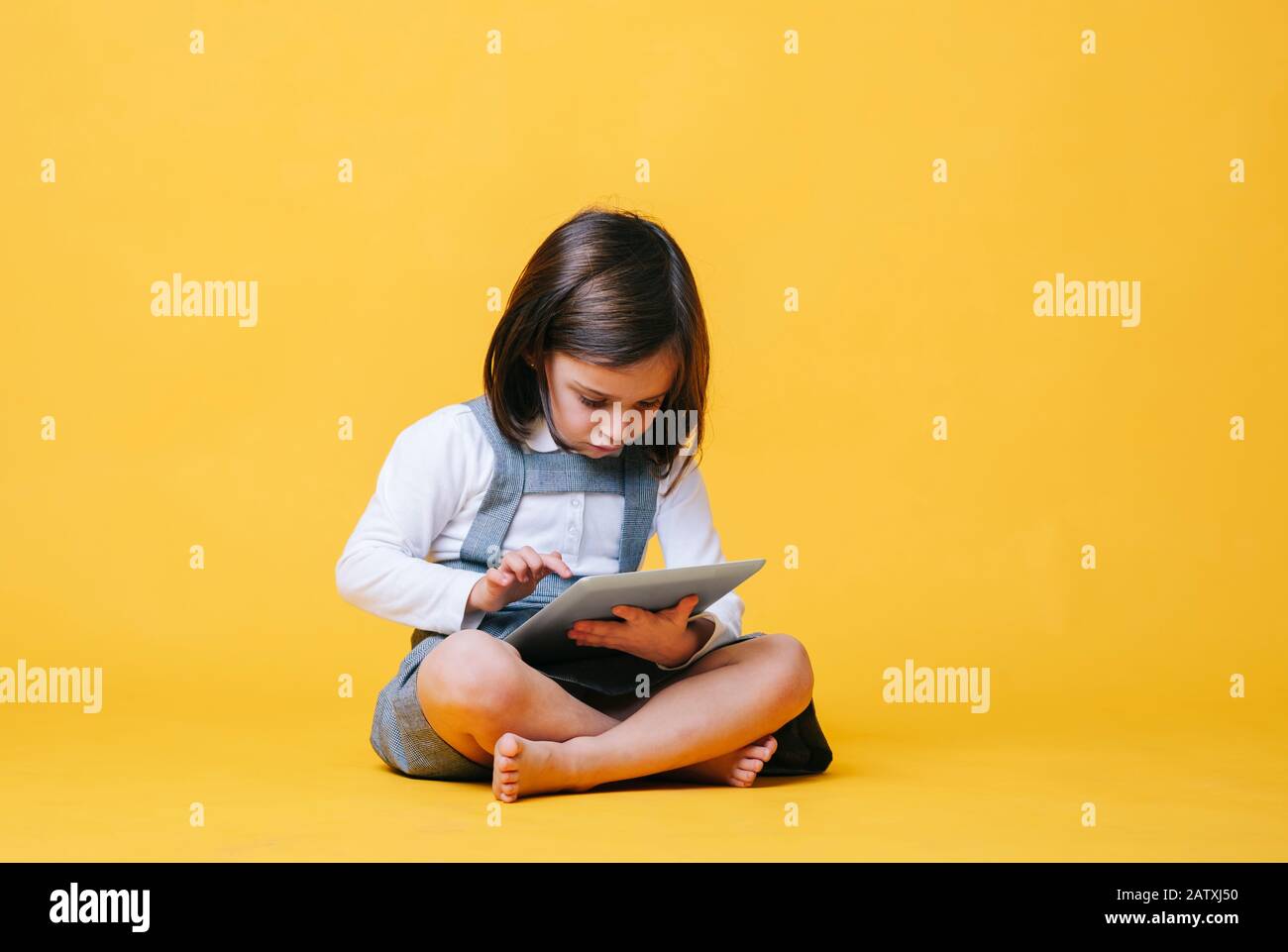 Ein kaukasisches Mädchen in einem grauen Kleid und einem weißen Hemd verwendet und spielt mit einer Tablette auf einem gelben Hintergrund Stockfoto