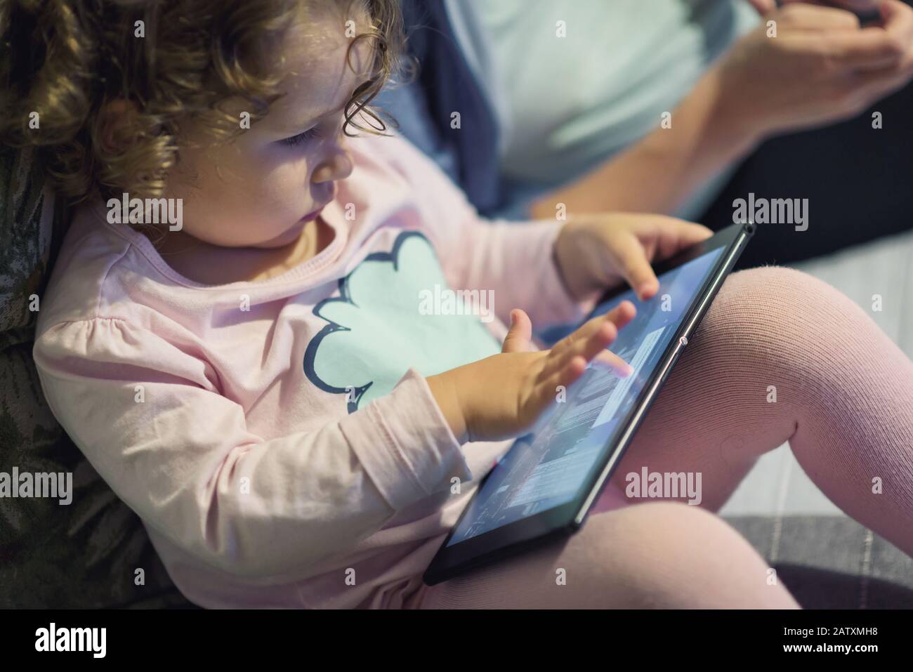 Kleines Mädchen sitzt in der Nähe ihrer Mutter und benutzt ein digitales Tablet. Zwei Jahre lang sieht ein Kind zu Hause ein elektronisches Gerät aus. Stockfoto