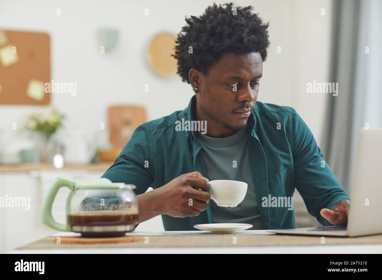 Junger Afrikaner, der zu Hause online arbeitet, hält eine Tasse Kaffee und tippt auf dem Laptop Stockfoto