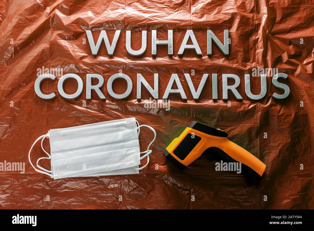 Worte wuhan Coronavirus mit Metallbuchstaben auf orange zerknitterter Kunststoff-Filmkulisse mit Gesichtsmasken und Infrarotthermometer - flach mit dram Stockfoto
