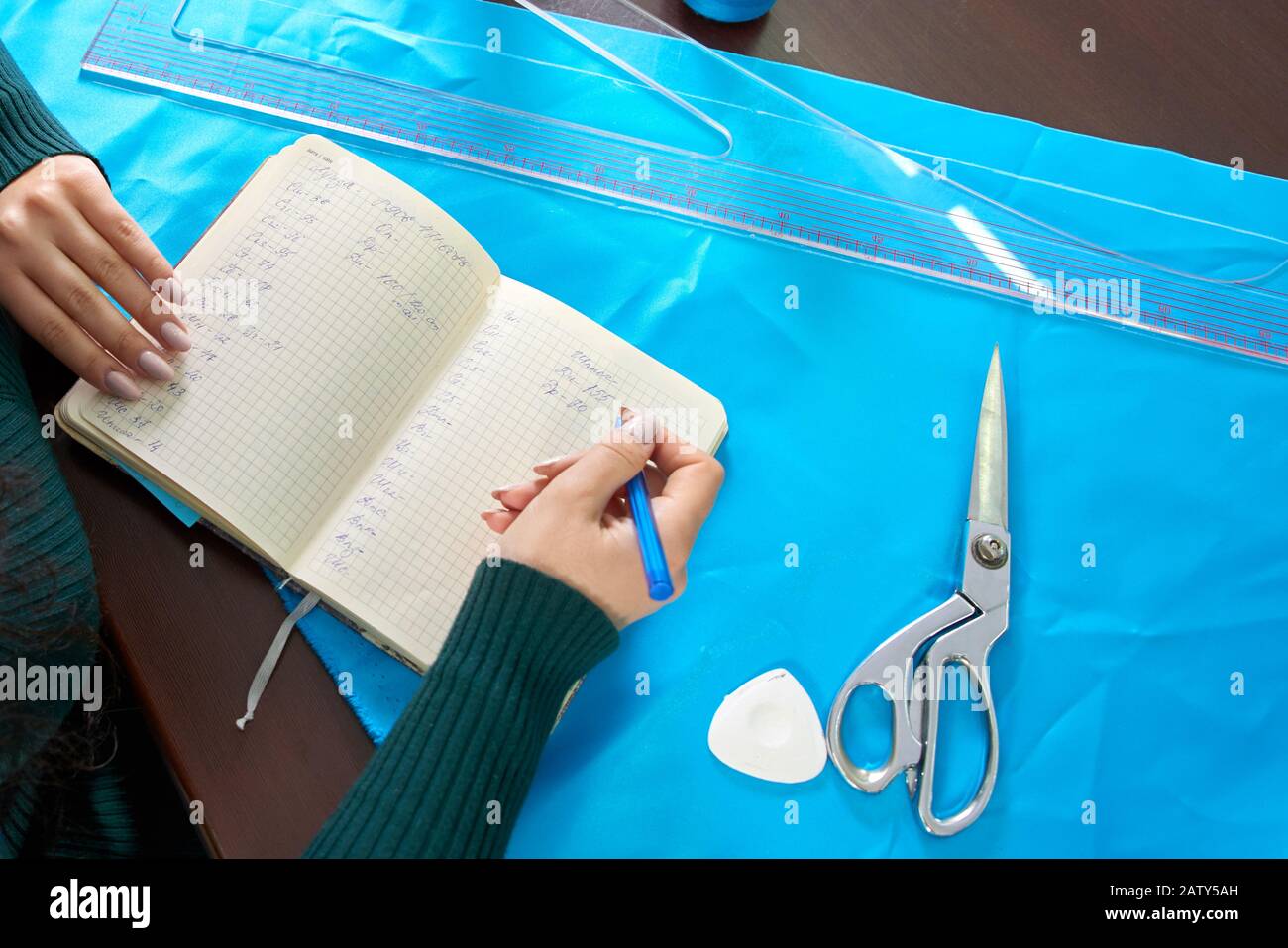 Schöne Näherin mit langen Haaren schreibt in einem Notizbuch. Der Schneider schafft eine Sammlung von Outfits. Junge Frau Designerkleidung notiert sich Ideen in Anm. Stockfoto
