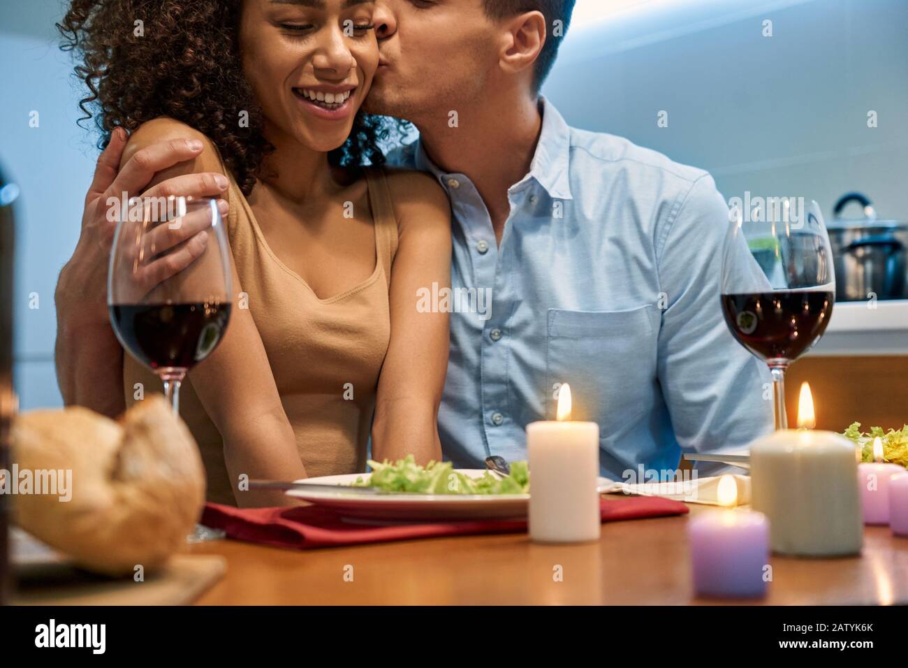 Junger Erwachsener, der eine afro-amerikanische Frau küsst und zu Hause ein romantisches Abendessen serviert Stockfoto