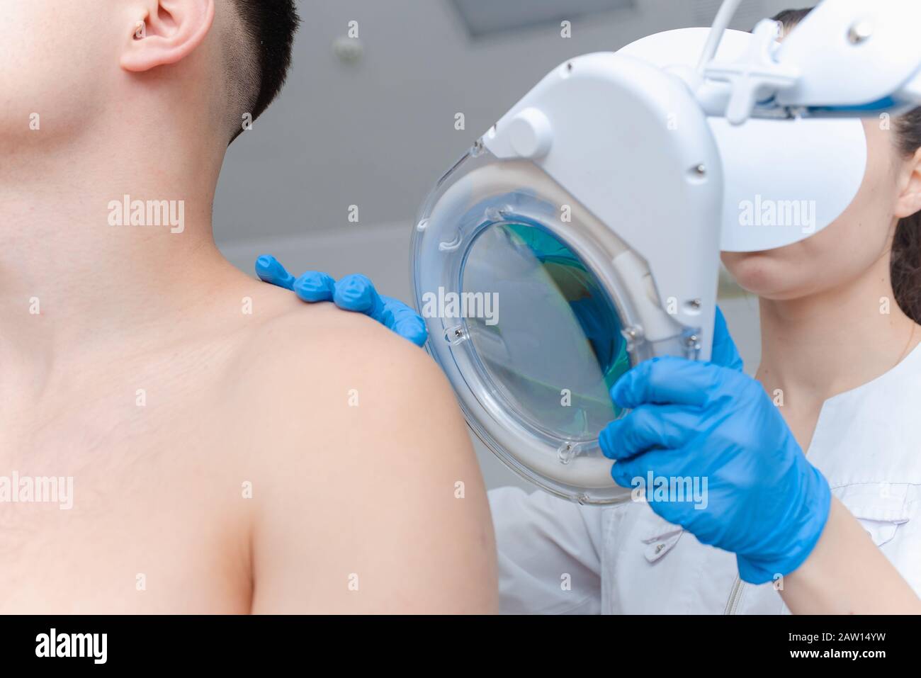 Ein Dermatologe untersucht die Haut am Körper eines jungen Mannes durch eine Lupe einer Kosmetologie-Lampe. Schönheitsbehandlungen Stockfoto