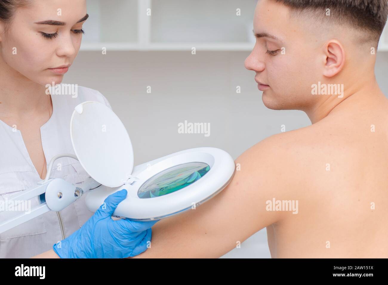 Ein Dermatologe untersucht die Haut am Körper eines jungen Mannes durch eine Lupe einer Kosmetologie-Lampe. Schönheitsbehandlungen Stockfoto