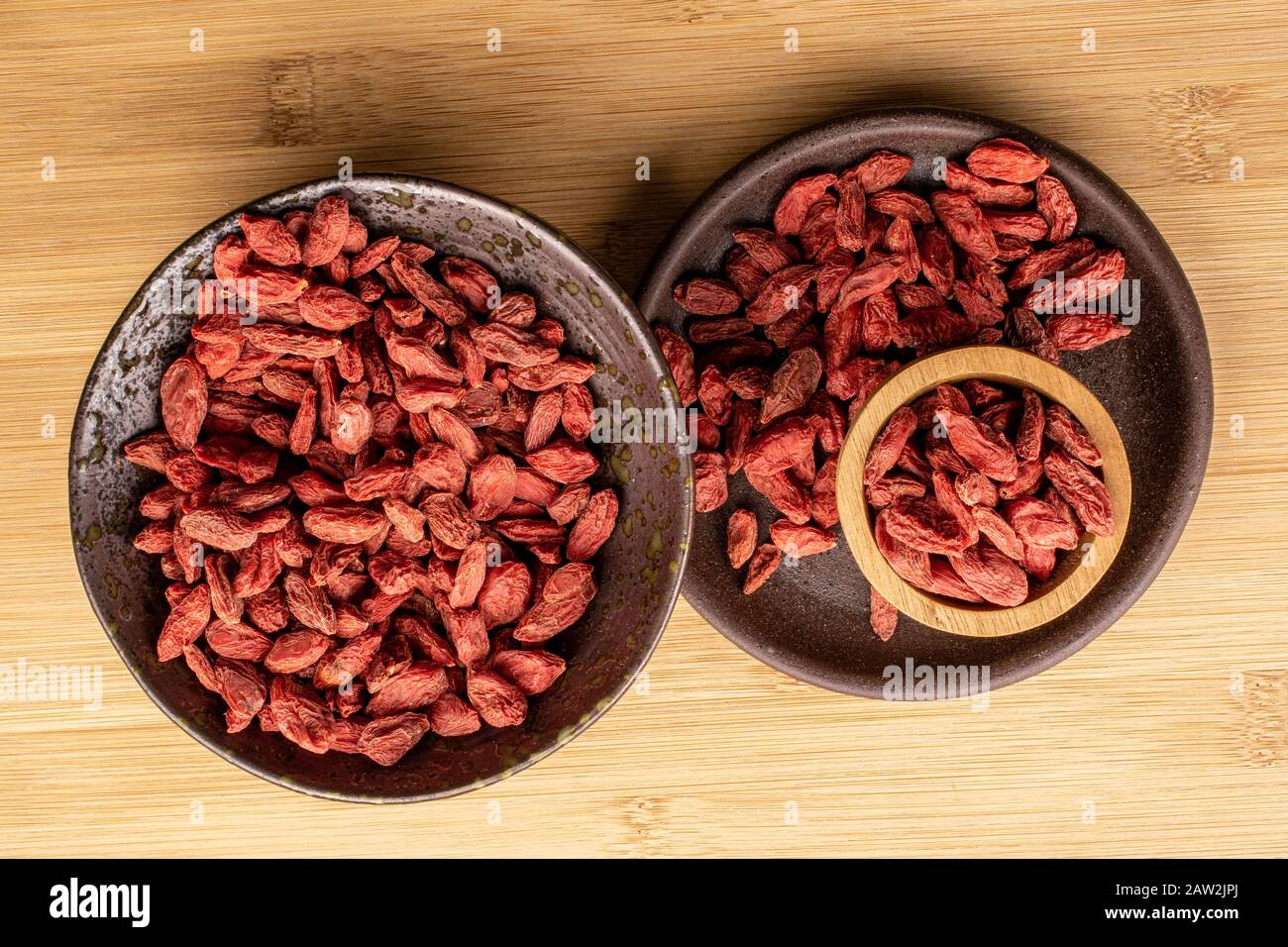 Viel getrockneter roter Goji in glasierter Schüssel mit brauner Keramikcoaster in Bambusschale auf hellem Holz flach Stockfoto