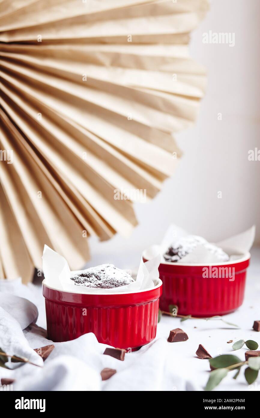 Schokoladenmuffins in roten Tassen. Kleines glasiertes Keramikramekin mit braunen Kuchen auf grauem und weißem Grund Stockfoto
