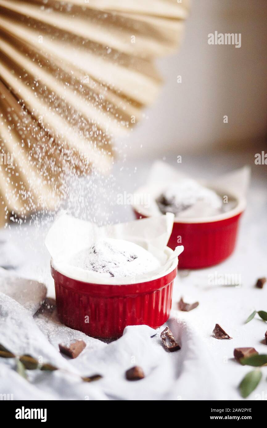Schokoladenmuffins in roten Tassen. Kleines glasiertes Keramikramekin mit braunen Kuchen auf grauem und weißem Grund Stockfoto