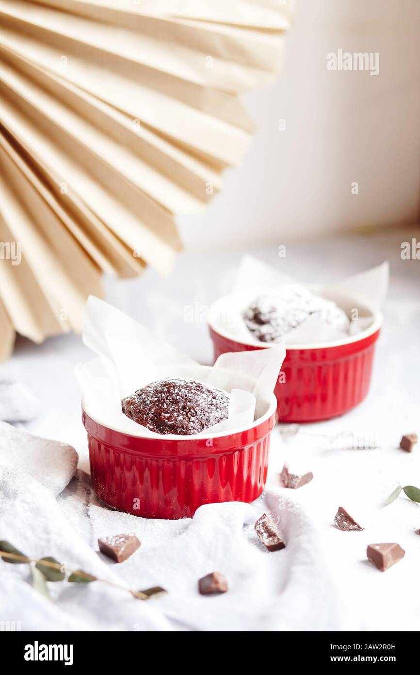 Schokoladenmuffins in roten Tassen. Kleines glasiertes Keramikramekin mit braunen Kuchen auf grauem und weißem Grund Stockfoto