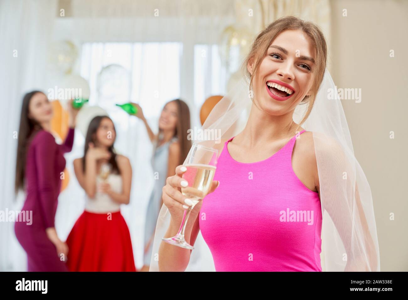 Vorderansicht der hübschen jungen lachenden Frau im Schleier, der morgens auf dem Bett sitzt und Glas mit Champagner hält. Schöne Frau, die sich auf der Junggesellen-Party lustig macht, Brautleute mit Flaschen hinterher. Stockfoto