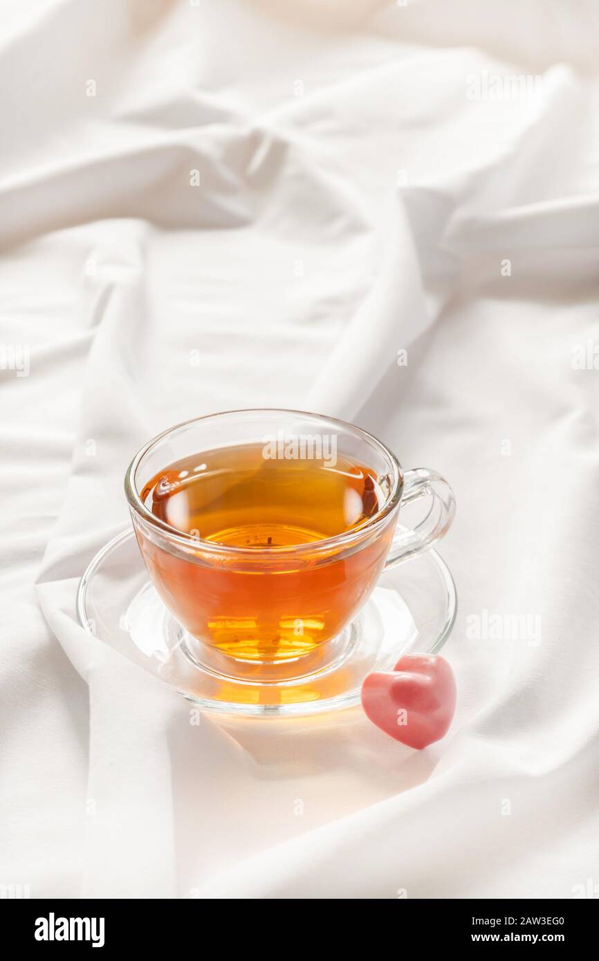 Romantisches Stimmungskonzept. Tasse Tee mit herzförmigem rosafarbenem Süßigkeiten auf weißem Bett mit Kopieraum. Stockfoto