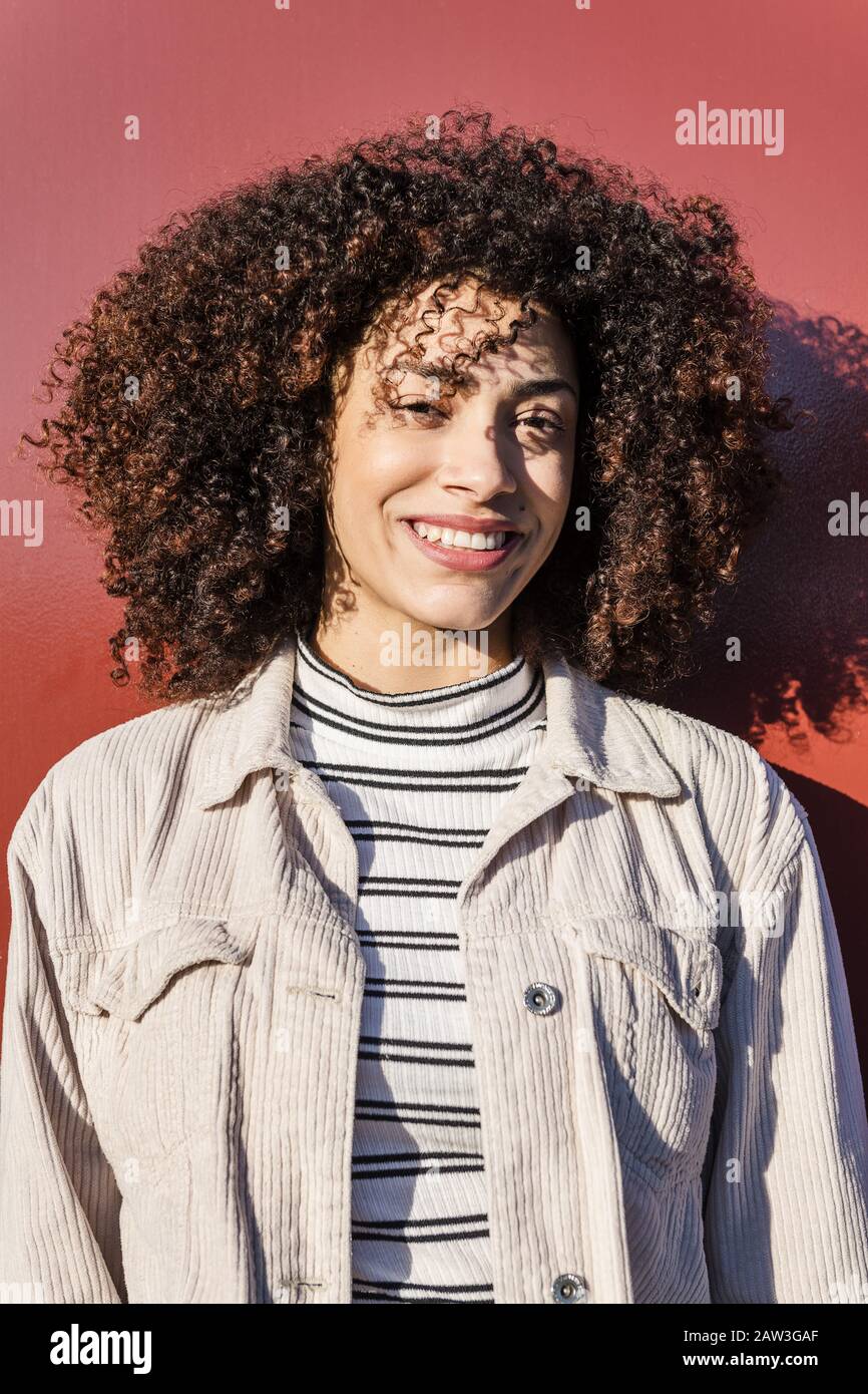 Vertikales Porträt einer schönen lächelnden Latina-Frau mit geschweiften Afro-Haaren auf rotem Hintergrund, Konzept der Schönheit und Vielfalt Stockfoto