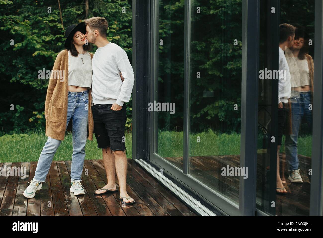 Stilvolles Hippie-Paar, das auf der Holzterrasse umarmt und küsst und sich in der Hütte in den Bergen entspannt. Fröhliche junge Familie in modernen Outfits, die sich auf Backgr umarmt Stockfoto