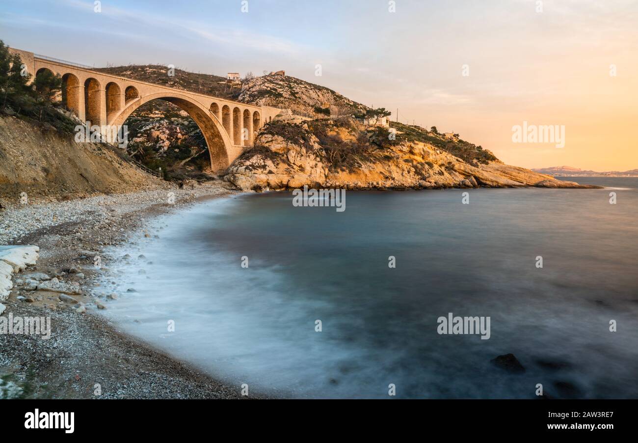 Lange Exposition des Sonnenuntergang in der Calanque des Eaux Salees oder des Salzwassereinlaufs mit Kieselstrand und alter Eisenbahnbrücke in der Nähe von Marseille France Stockfoto