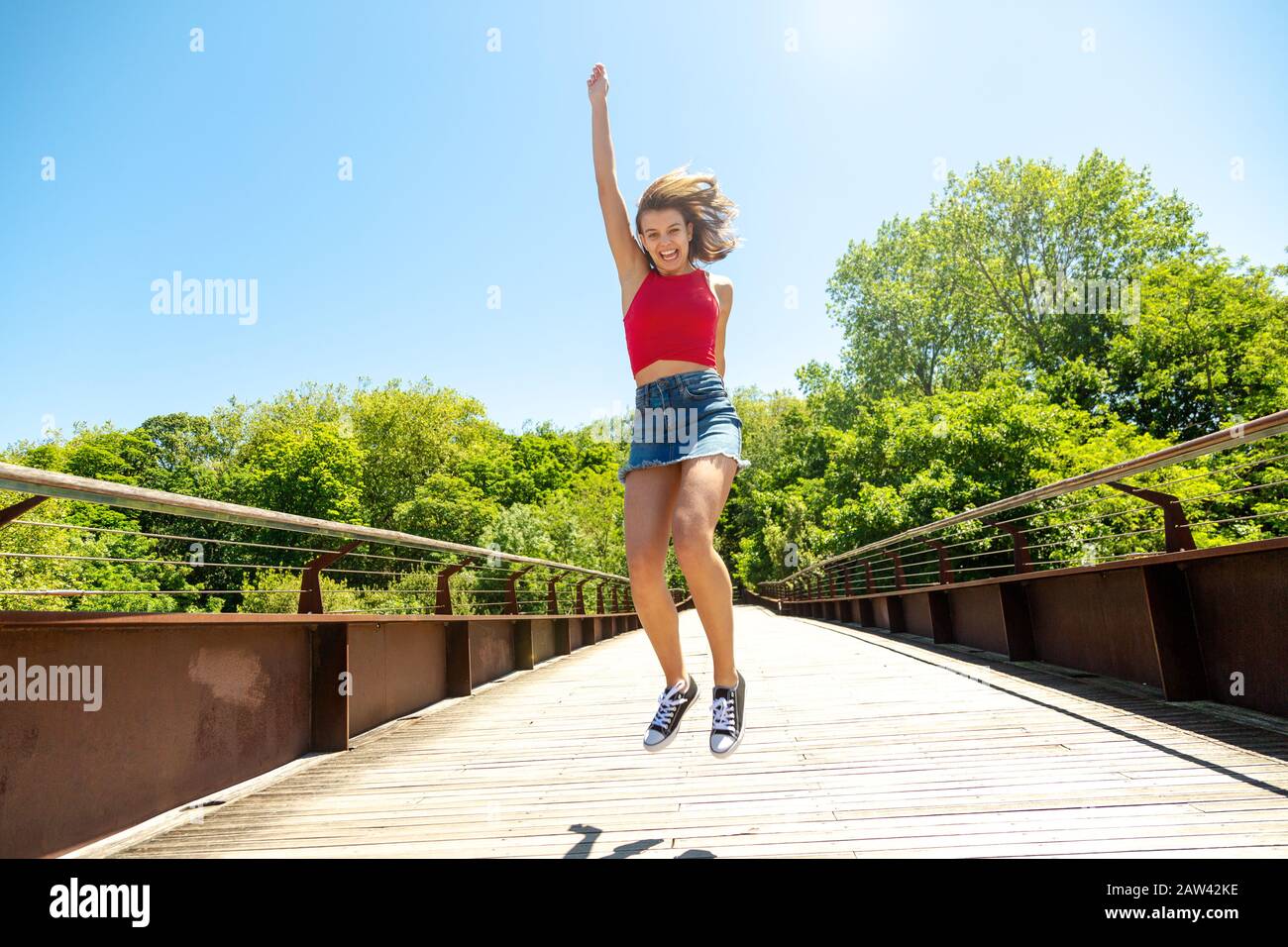 Fröhliche junge schöne Frau im roten Oberteil und Minirock, die an einem sonnigen Tag auf einer Brücke springt Stockfoto
