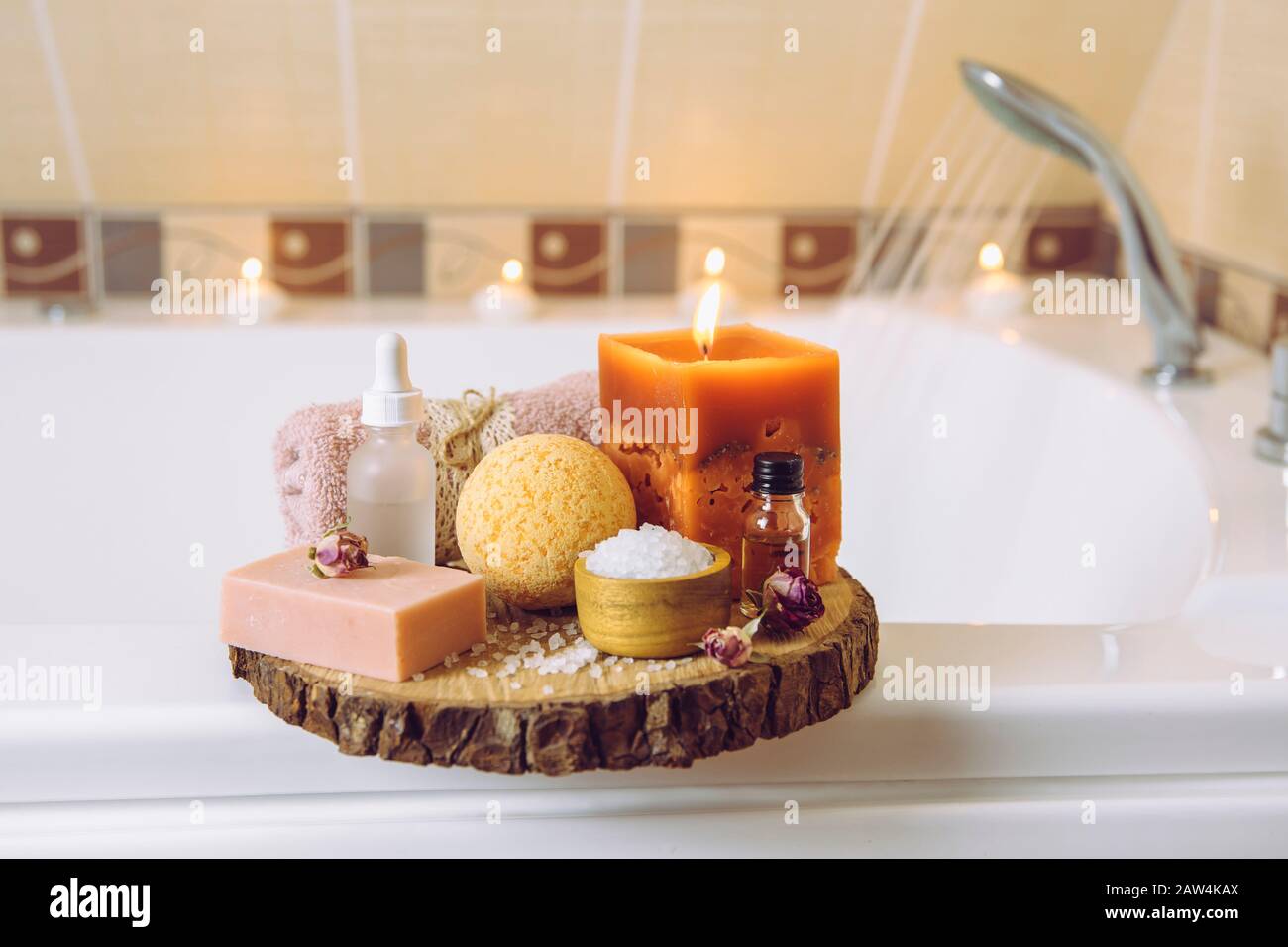 Home Spa-Produkte auf Holz-Disc-Tablett: Seifenbar, Badbombe, Aromabadsalz, ätherische und Massageöle, Kerze brennen, Handtuch im Badezimmer. Stockfoto