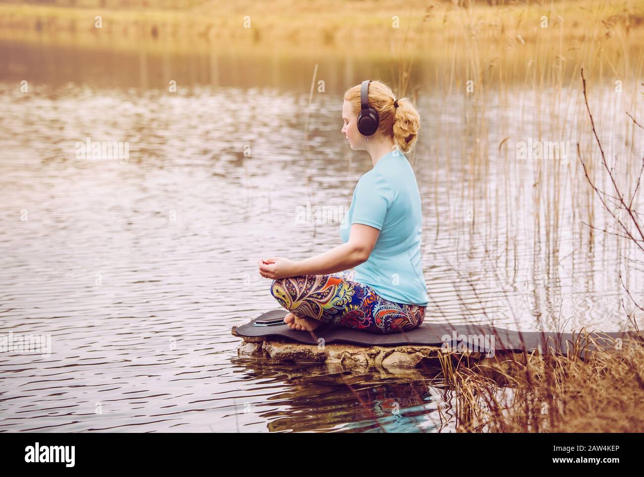 Blonde Haarfrau sitzt draußen auf dem Wasser und hört die Audio-geführte Meditation vom Smartphone mit blauer T-Shirt Sportbekleidung. Warmer sonniger Tag. Stockfoto