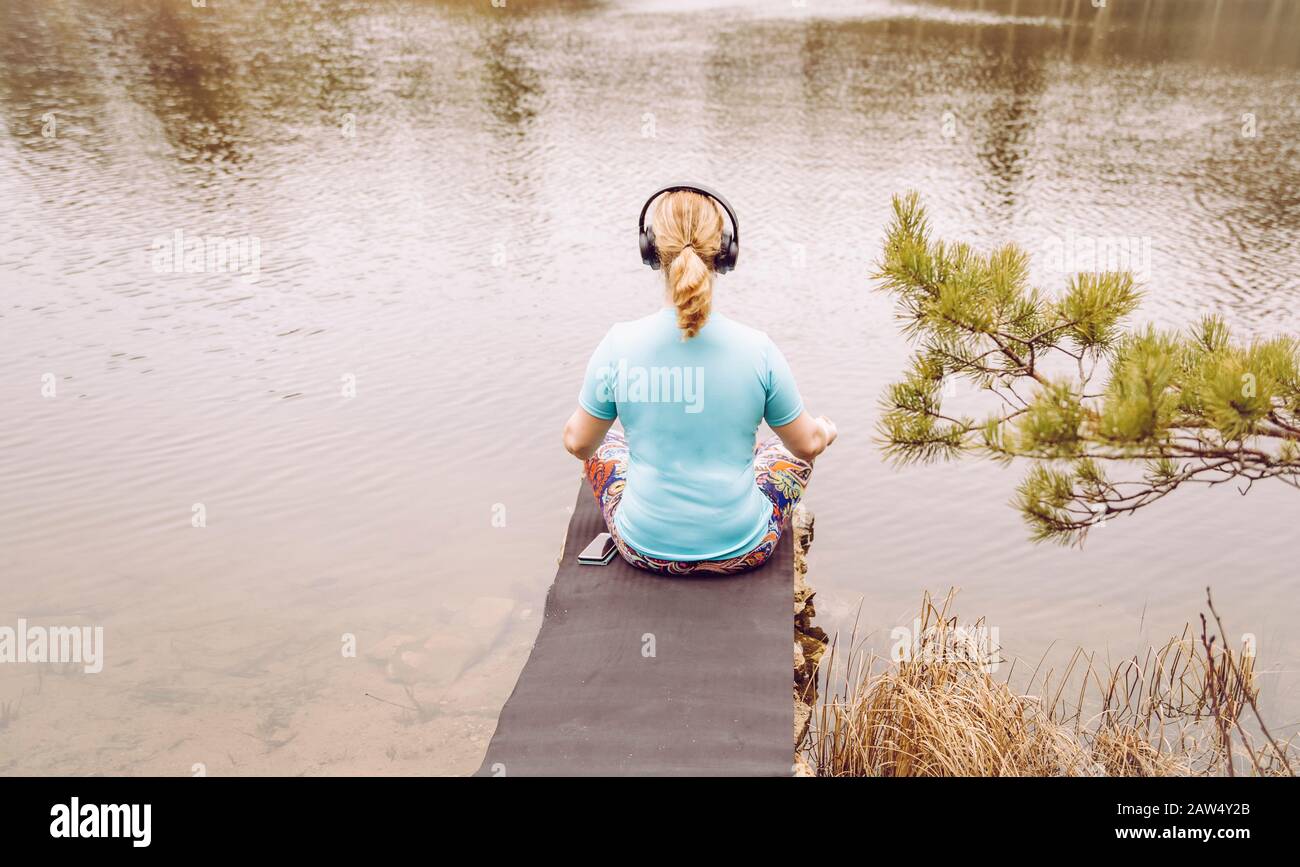 Blonde Haarfrau sitzt draußen auf dem Wasser und hört die Audio-geführte Meditation vom Smartphone mit blauer T-Shirt Sportbekleidung. Warmer sonniger Tag. Stockfoto