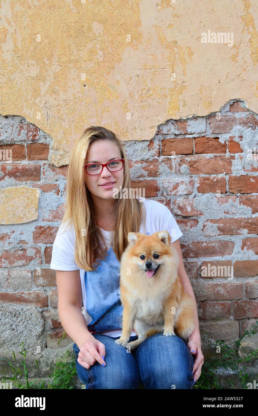 Junges, süßes Mädchen nebenan, das rote Brille, Jeans und T-Shirt trägt und mit ihrem Haustier vor der Grunge Ziegelwand posiert Stockfoto