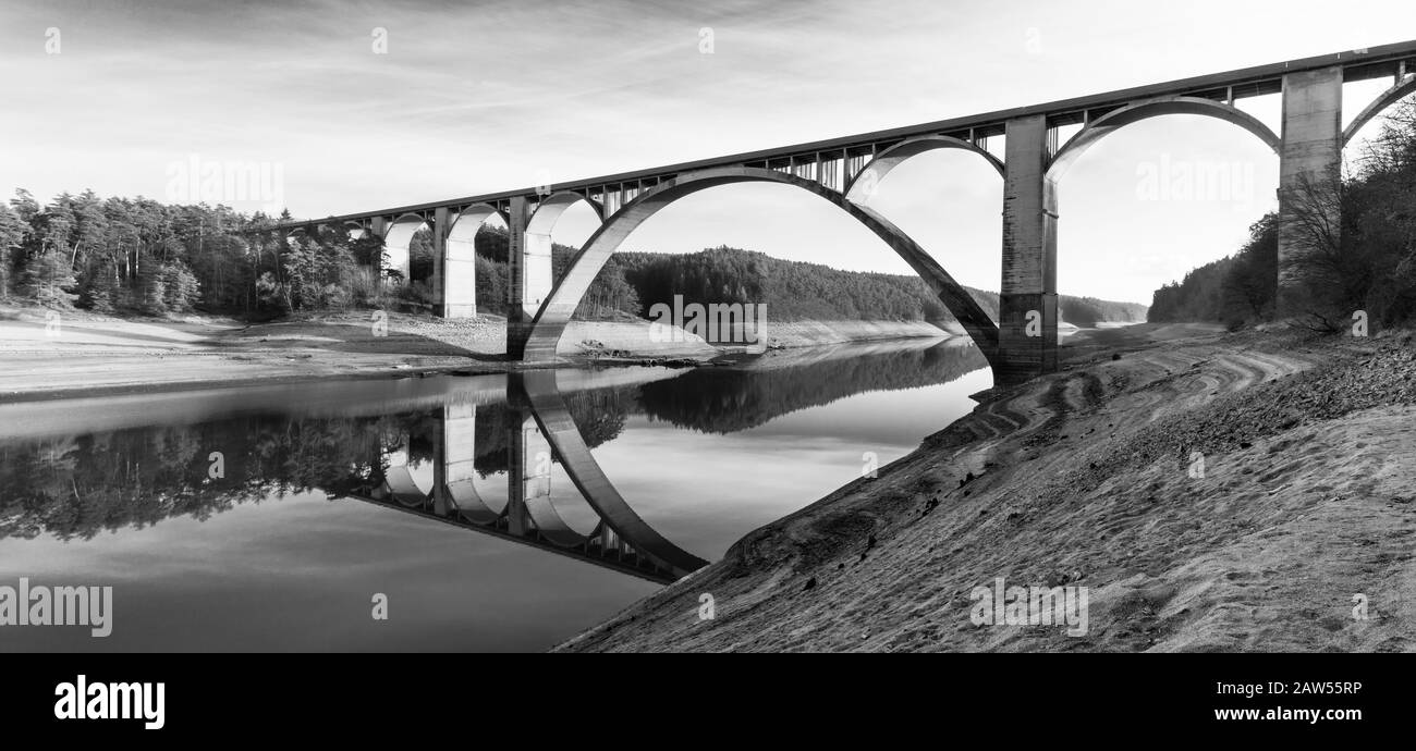 Hoher Bogenstraßenviadukt über dem Orlik-Stausee. Panoramische Schwarzweißansicht. Podolsko-Brücke, Tschechien. Schlammige Bank und Spiegelung in der Wasseroberfläche. Stockfoto