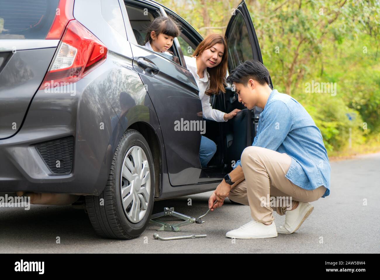 Junger asiatischer Vater wechselt den Reifenpannen auf seinem Auto und löst die Muttern mit einem Radschlüssel, bevor er Fahrzeug und Mutter aufbocken kann Stockfoto